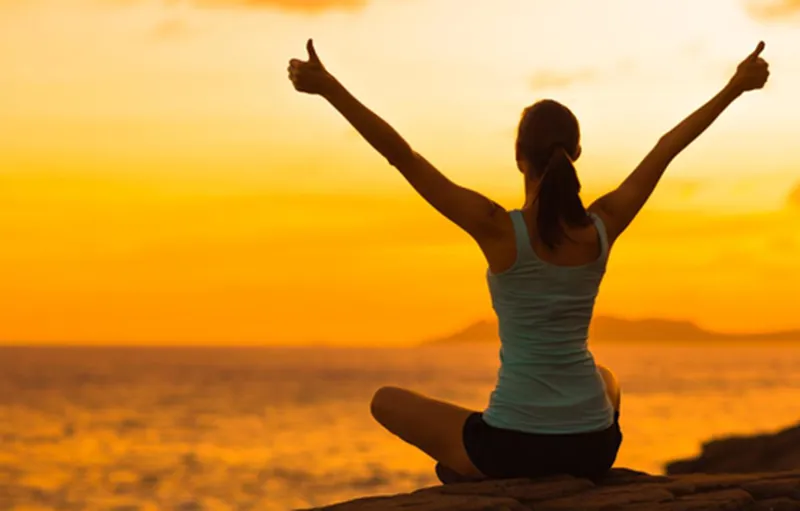 Woman sitting by the sea at sunset with her arms raised